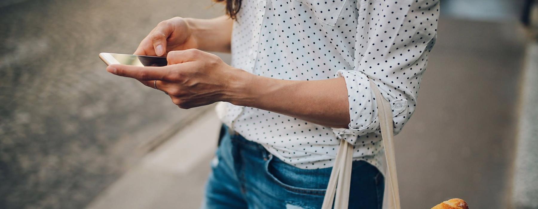 woman walks down the street and texts with a bag of groceries on her arm